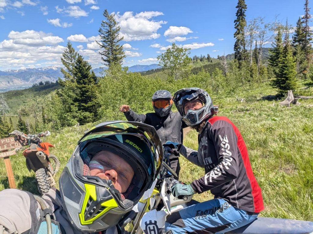 Three riders in helmets grinning on a high mountain ridge in the Idaho backcountry, dirt bikes beside them